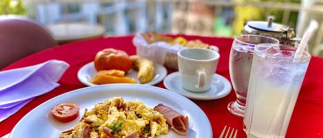 Una mesa de comedor con un plato de huevos revueltos, un panecillo y un tomate. También hay dos vasos de té helado y una taza blanca sobre la mesa.