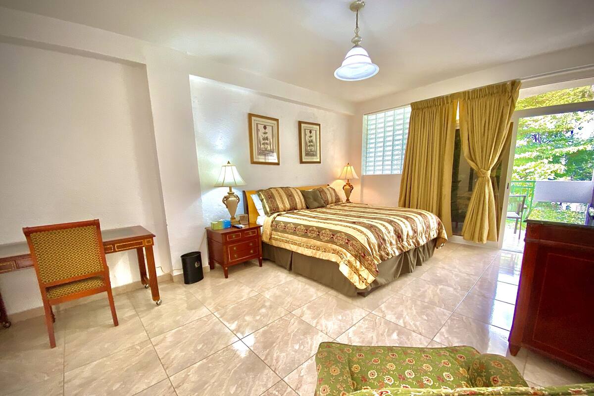 A bedroom with a bed, a chair, a nightstand, a lamp, a window with curtains, and a view of greenery outside.