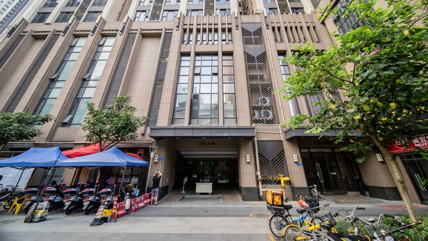 Un edificio moderno con una entrada destacada, varios pisos y un árbol en frente.