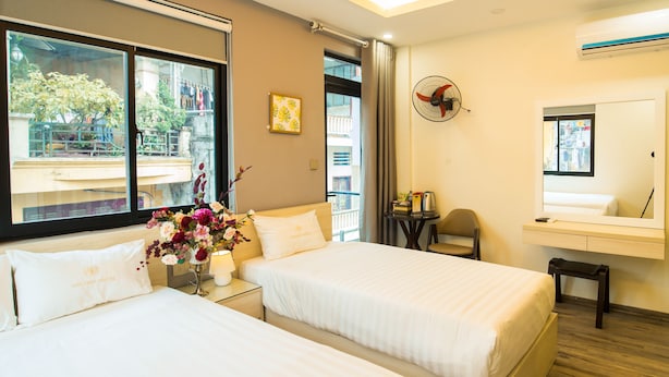 A hotel room with two beds, a small table with a chair, a window with a view of the outside, and a wall-mounted fan. - Golden hotel (Hanoi)