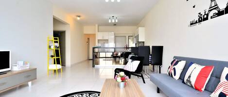 A modern living room with a sofa, coffee table, and TV. There is a kitchen in the background with cabinets and a dining area.