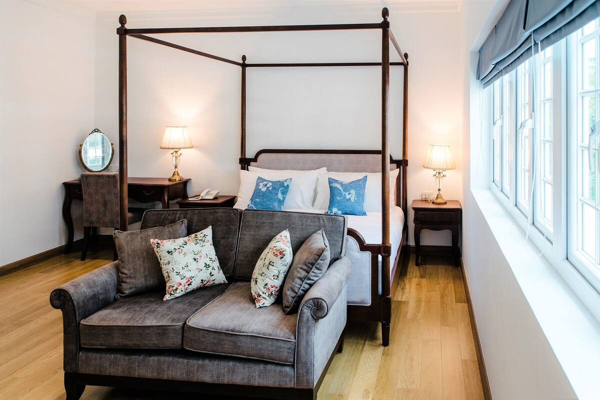 Ein Bett mit Baldachin, eine graue Polstersofa und florale Kissen in einem Zimmer mit Holzboden und einem großen Fenster.