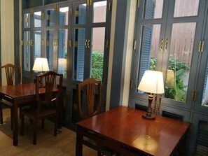 A dining area with wooden tables and chairs, two lamps, and a view of greenery through blue-framed windows. - Baan Vajra (Bangkok)