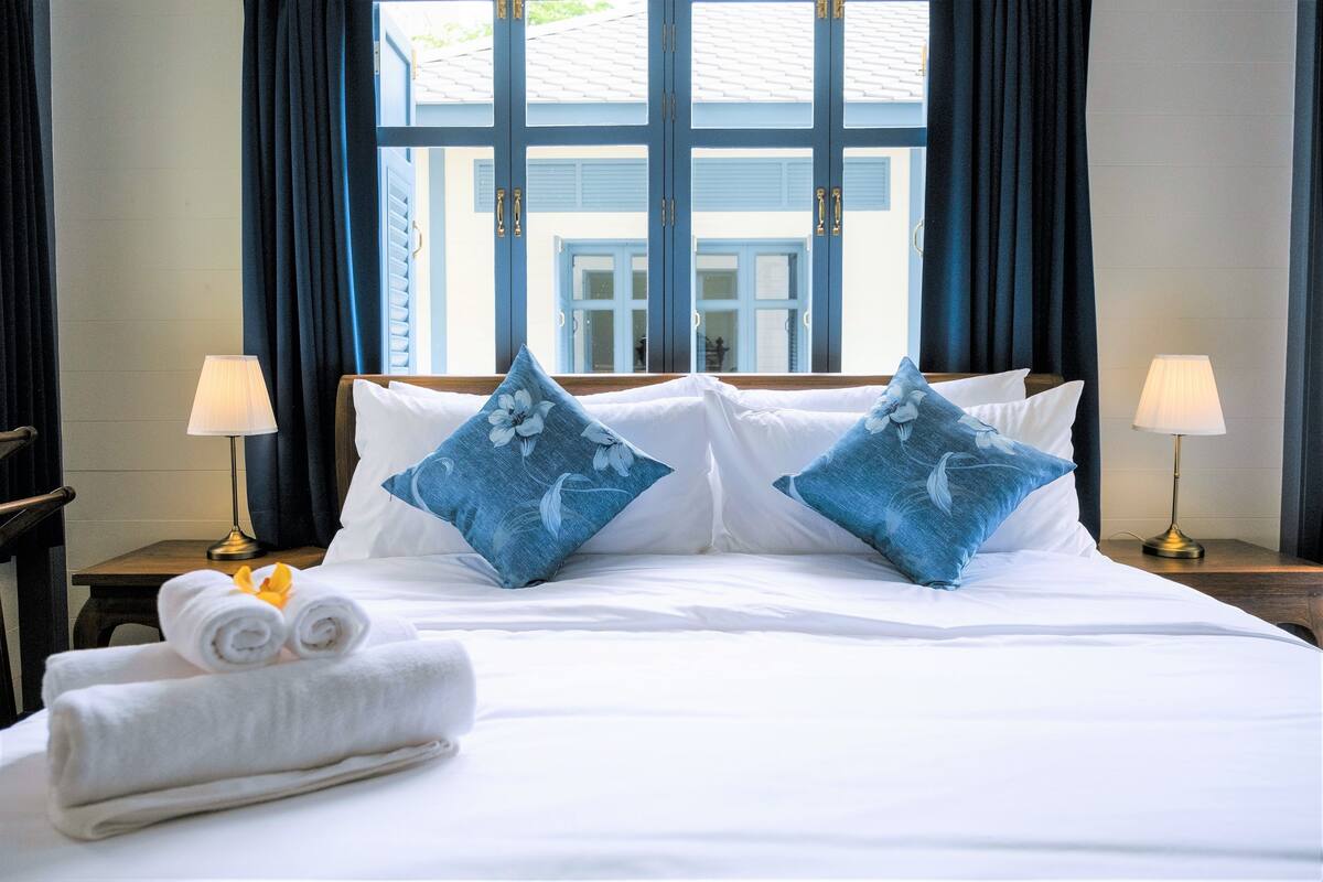 Ein ordentlich bezogenes Bett mit weißen Leinen und blumigen blauen Kissen, ein aufgerolltes Handtuch und ein kleiner Tisch mit einer Lampe.