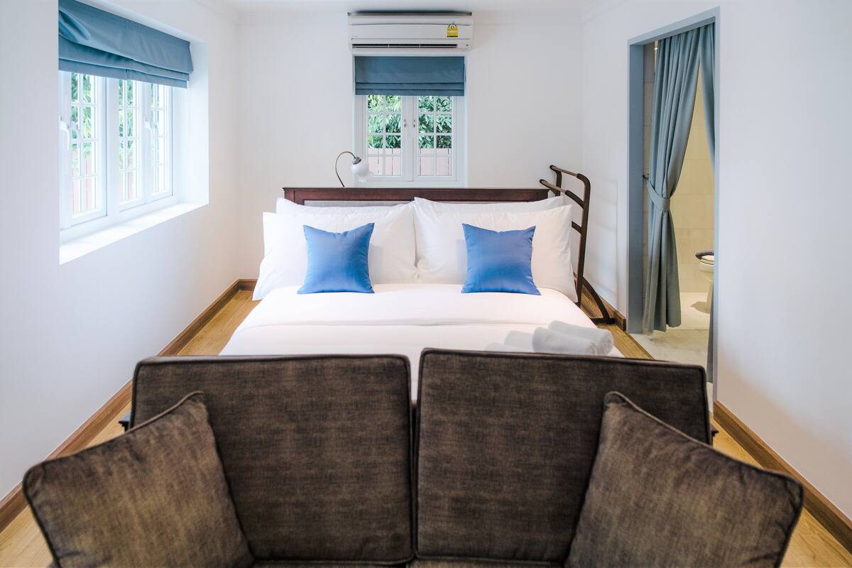 Ein Hotelzimmer mit einem großen Bett, zwei Kissen, einem Nachttisch, einer Lampe, einem Fenster mit Vorhängen und Blick auf Grünflächen.