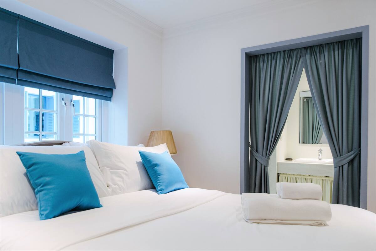 Ein Hotelzimmer mit Bett, Fenster mit Jalousien, Nachttischlampe und sichtbarem Badezimmer im Spiegel.