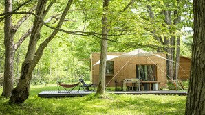 Una cabaña de madera en un bosque con una hamaca, una mesa y sillas afuera.