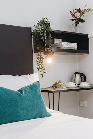A neatly arranged hotel room with a bed, a bedside table with a kettle, a small shelf with books, and a hanging plant.