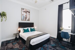 Hotellihuone, jossa on sänky, työpöytä, tuoli, kasvi ja taulu seinällä.