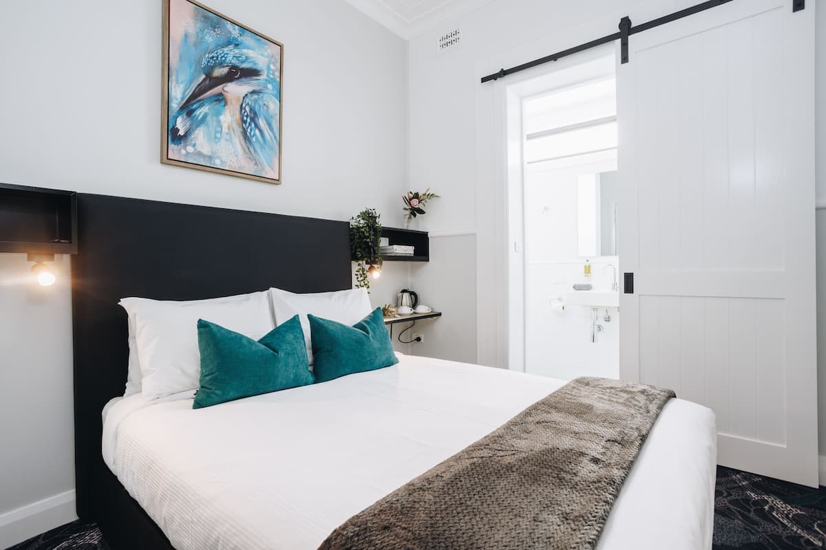 A modern hotel room with a large bed, a black headboard, teal pillows, a framed artwork, a small plant, and a nightstand with a lamp.