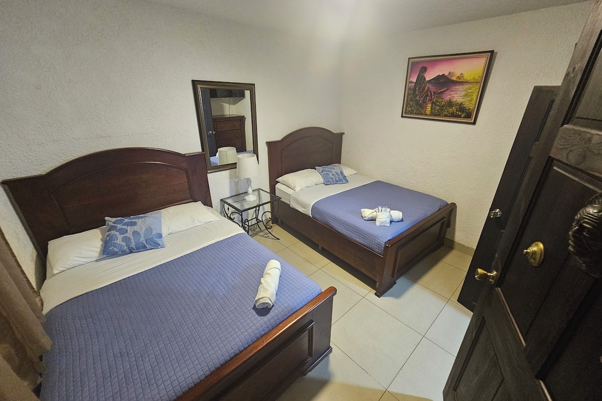 A hotel room with two beds, a small table, a mirror, and a painting on the wall.