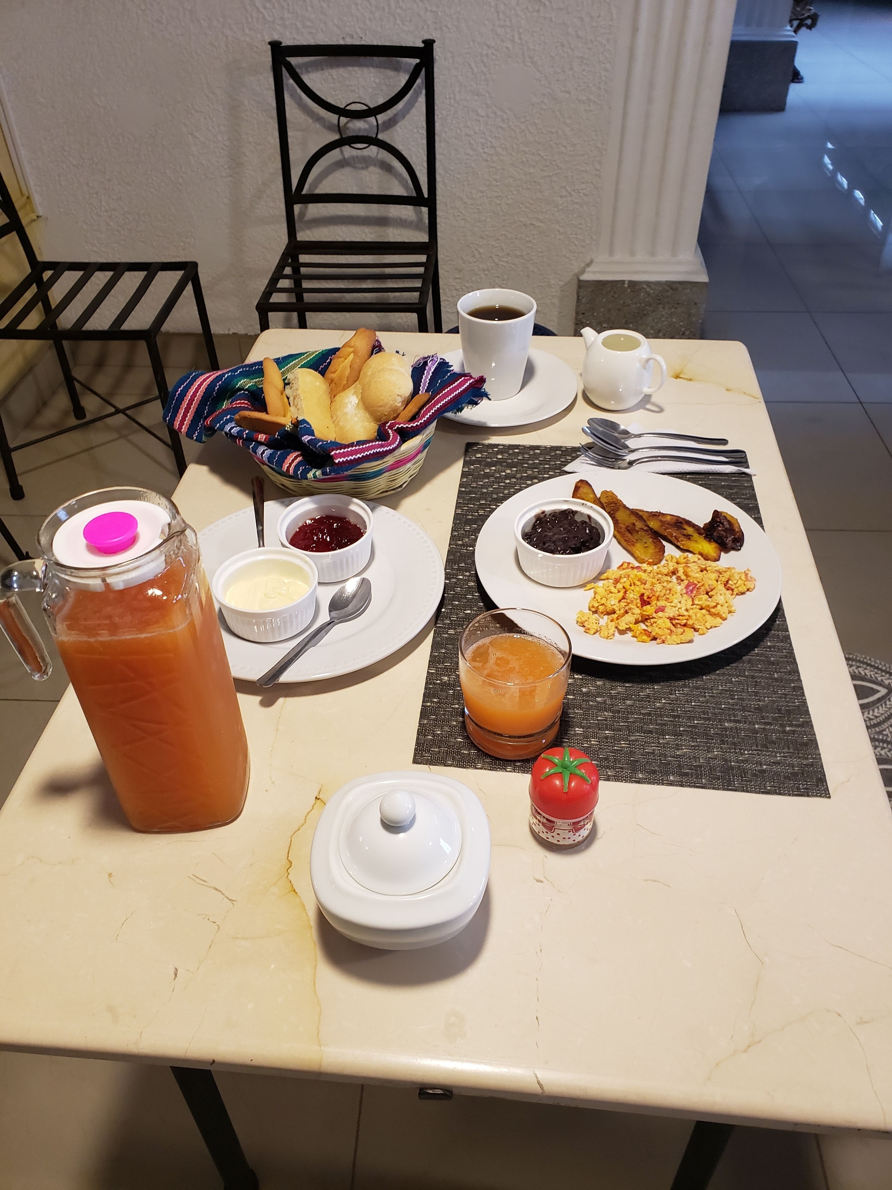 Una mesa de desayuno con una jarra de jugo de naranja, un bol de pan, una taza de café, un plato de huevos revueltos y un vaso de jugo de naranja.