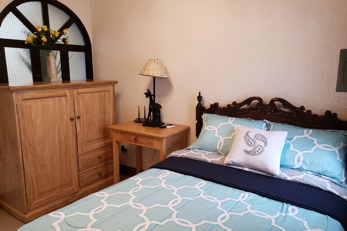 A bedroom with a bed, a wooden wardrobe, a nightstand with a lamp, and a window with a view.