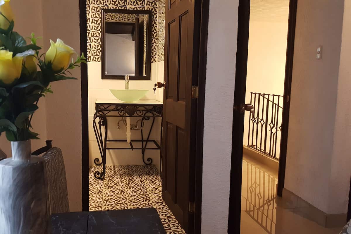 A narrow hallway with patterned floor tiles, a sink with a geometric mirror above it, and a vase with yellow flowers on a pedestal.