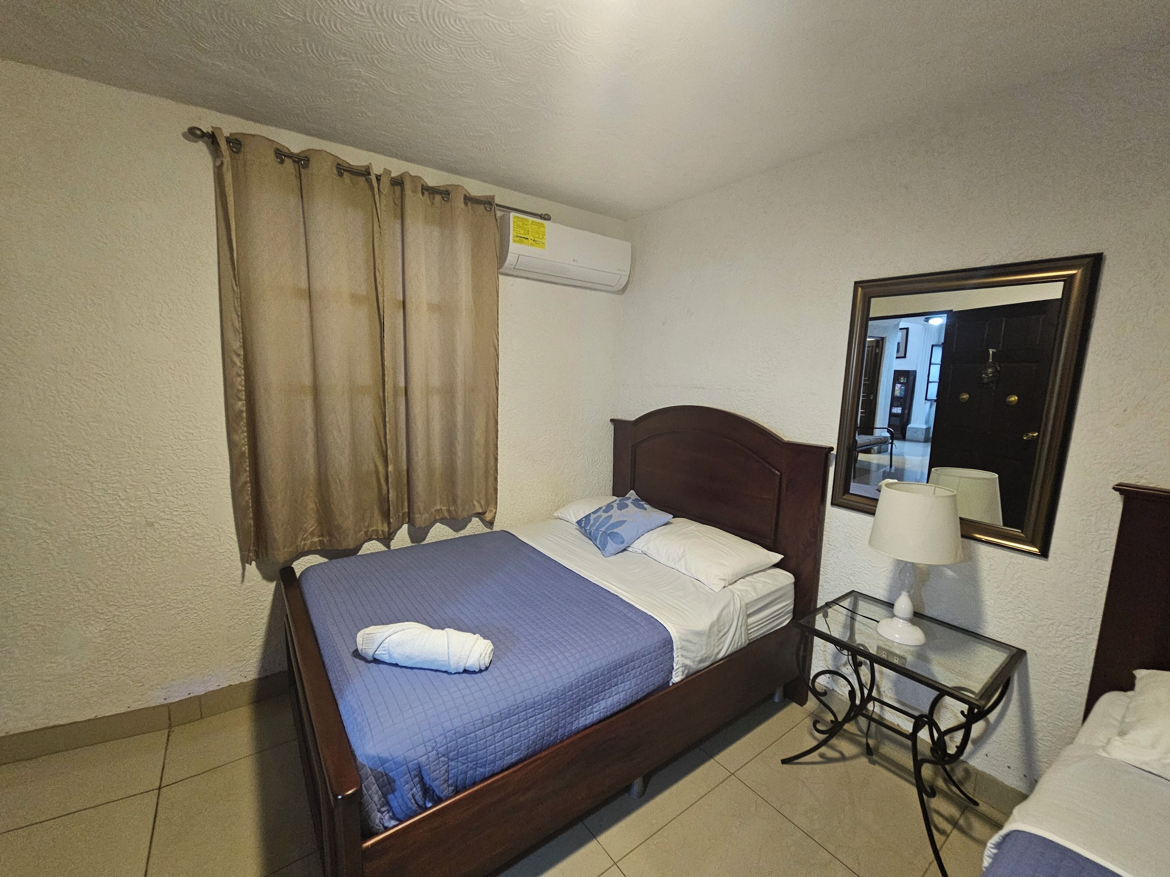 Un dormitorio con cama, cortinas, aire acondicionado, mesita de noche con lámpara y espejo.