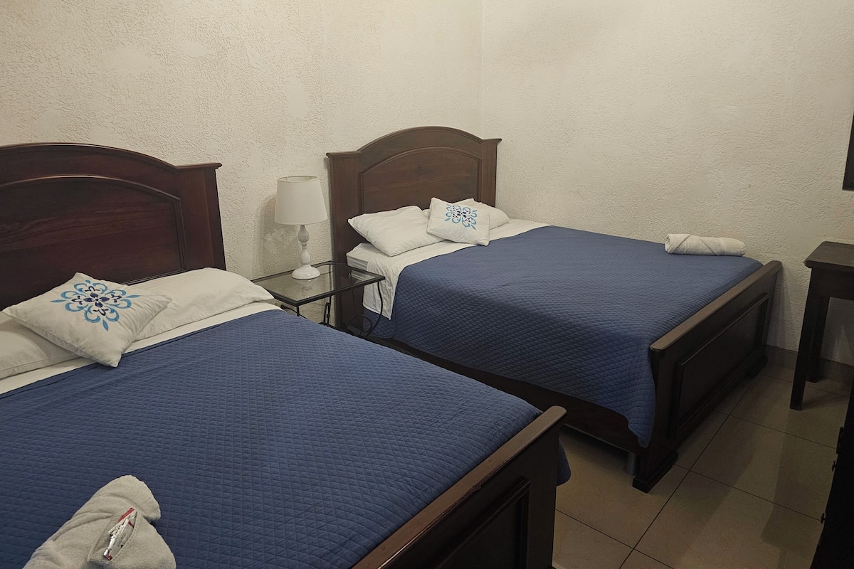 Two single beds with blue bedding, a bedside table with a lamp, and a white wall.