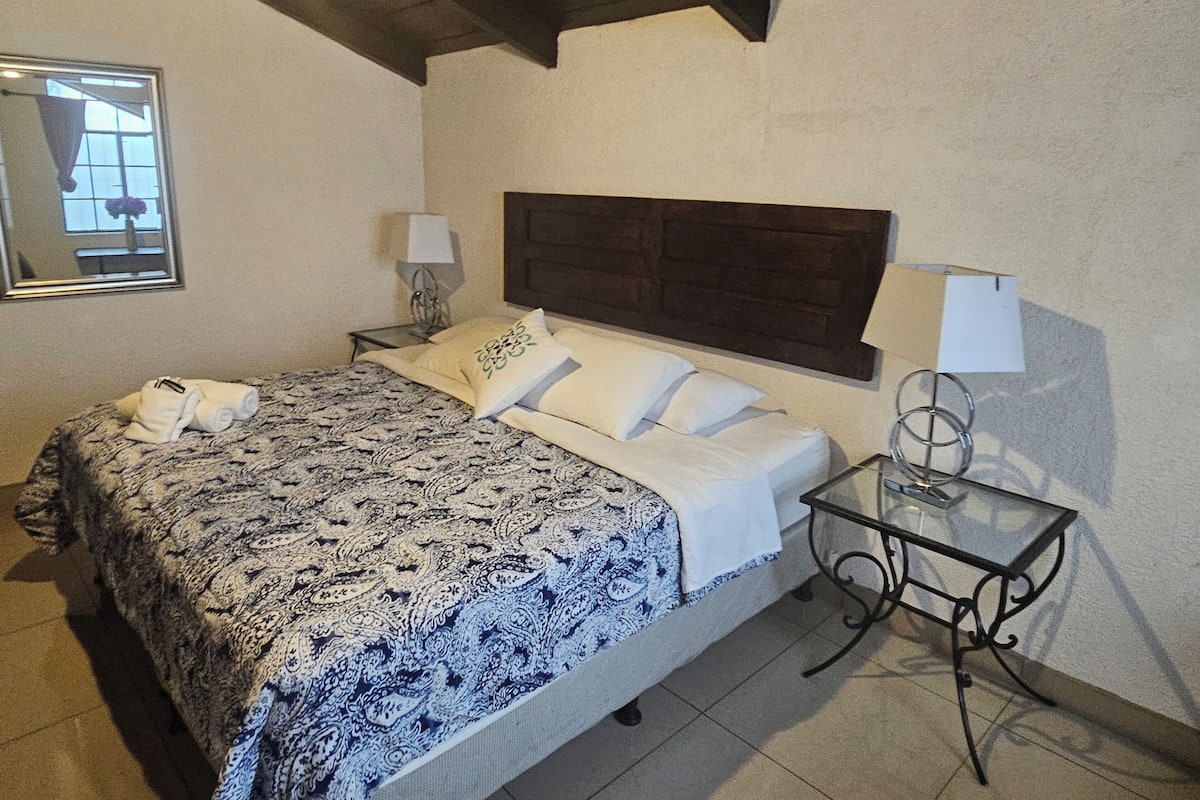 A bedroom with a bed, a nightstand, a lamp, and a glass table.