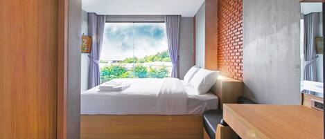 A modern hotel room with a bed, a wooden desk, and a view of greenery outside.
