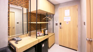 Habitación de hotel con un escritorio de madera, un teléfono, una tetera y una cafetera. Hay un espejo en la pared y una puerta con un cartel.
