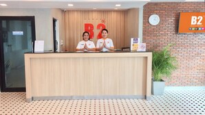 Un mostrador de recepción con dos empleados, un reloj y un cartel de B2 Boutique & Budget en la pared.