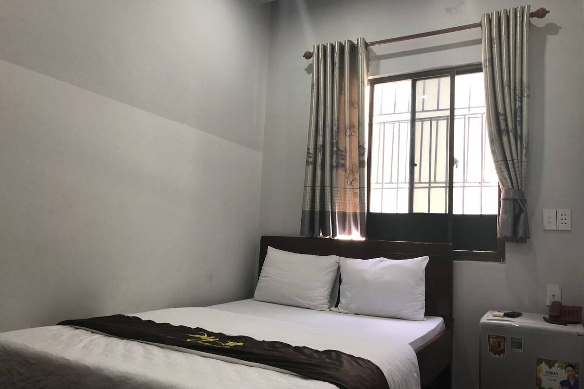 A single bed with white bedding and a dark brown blanket, a small bedside table with a phone and a water dispenser, and a window with curtains.