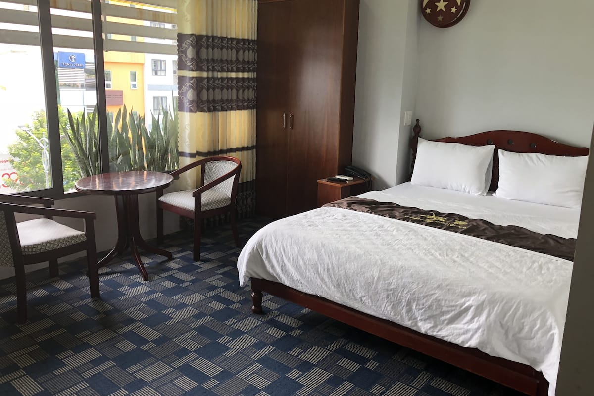 A hotel room with a bed, a wooden wardrobe, a round table with a chair, and a view of the outside through large windows.
