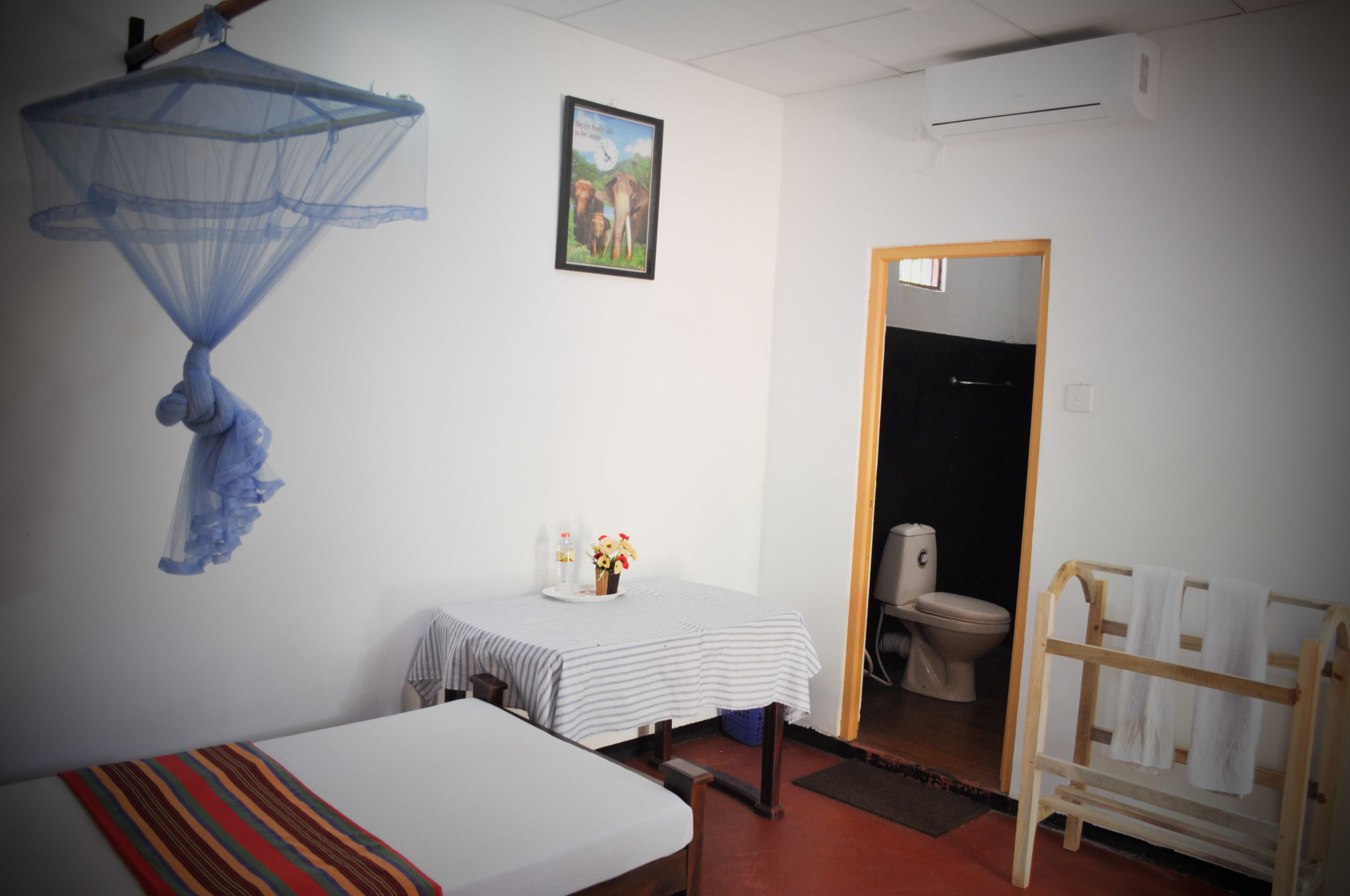 A room with a bed, a small table with a vase of flowers, a mosquito net, a framed picture, a wooden rack with towels, and a toilet visible through a doorway.