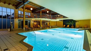 Una piscina interior con suelo de baldosas, un balcón con sillas y una escalera que lleva a un nivel superior.