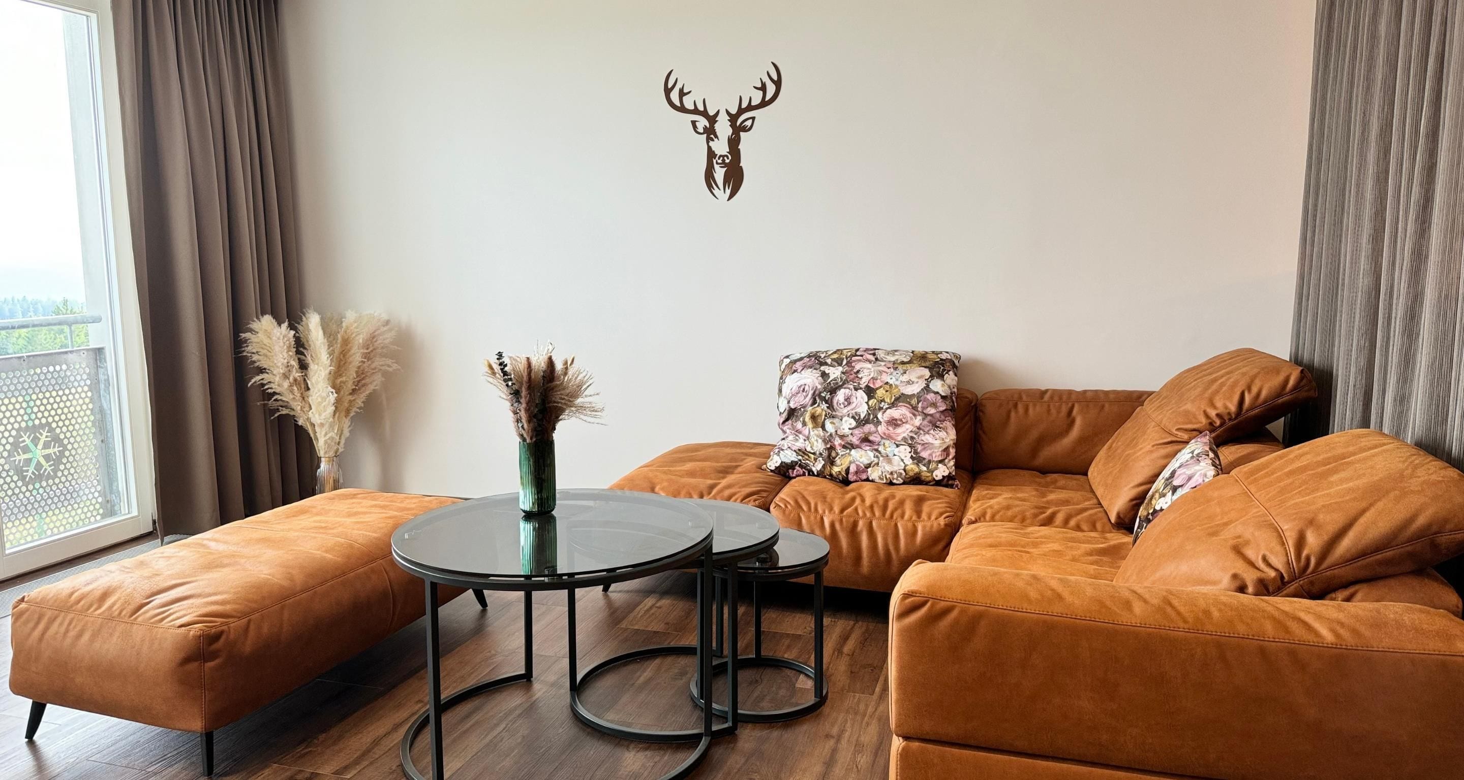 Una sala de estar con un sofá de cuero marrón, una mesa de centro redonda de vidrio y una cabeza de venado decorativa en la pared.
