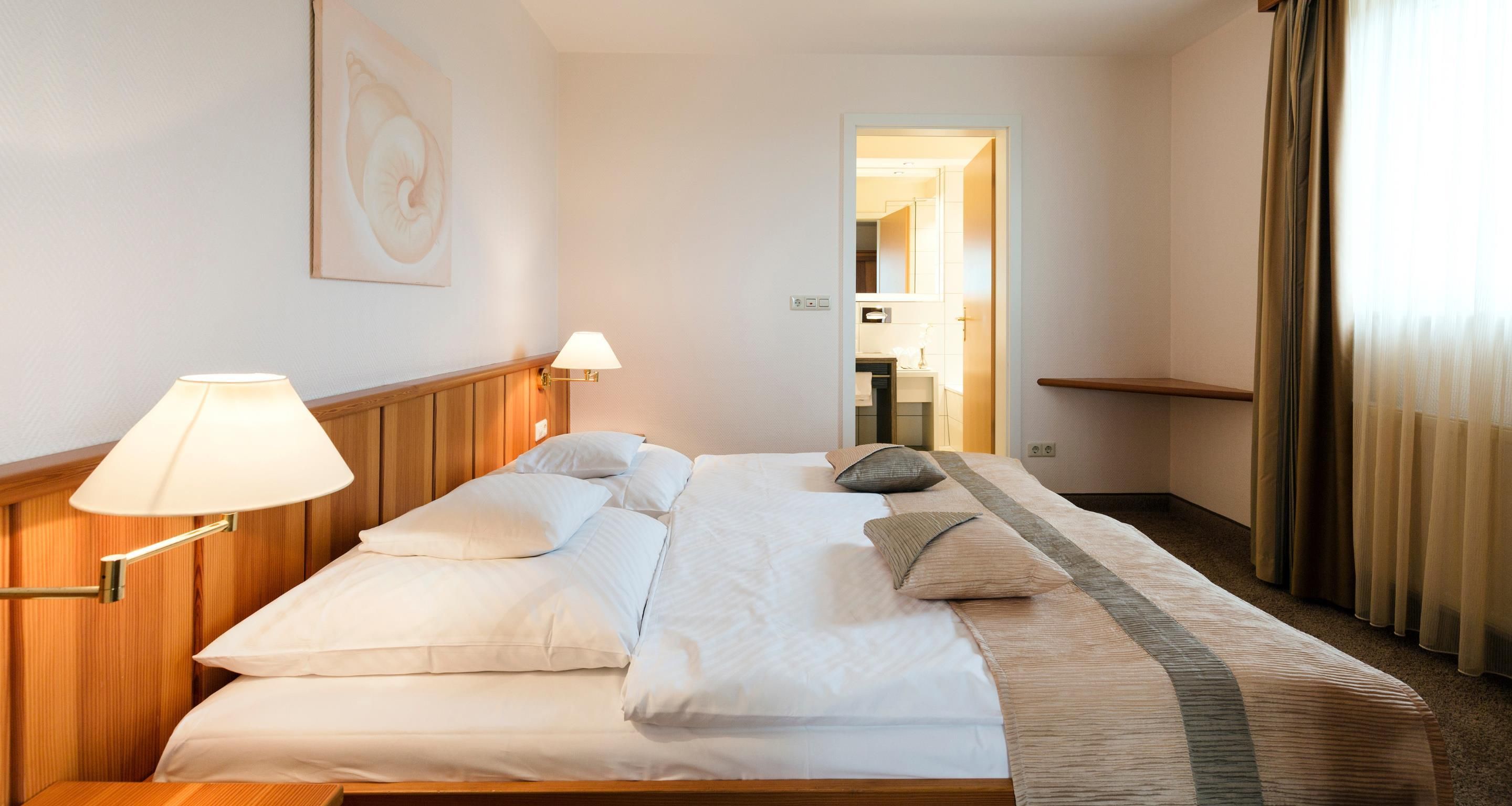 A hotel room with a bed, two bedside lamps, a headboard, a window with curtains, and a door leading to a bathroom.