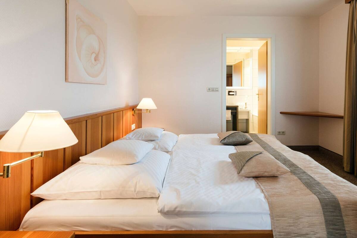 Une chambre d’hôtel avec un lit soigneusement fait, deux lampes de chevet, une tête de lit en bois, un tableau au mur et une vue sur la salle de bain par une porte ouverte.