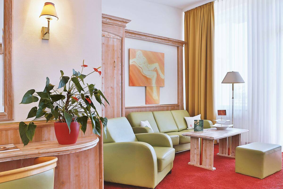 Une chambre d’hôtel avec un canapé vert, une plante en pot rouge, une table d’appoint en bois, une petite table blanche et un tapis rouge.