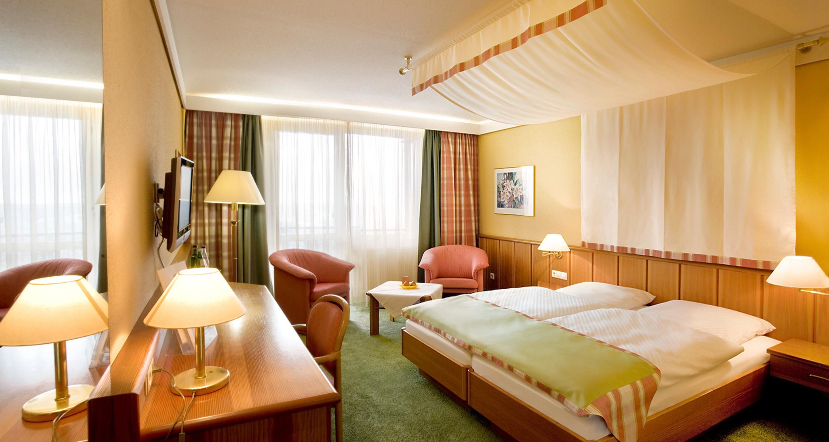 A hotel room with a bed, two chairs, a desk with a lamp, a TV, and a window with curtains.