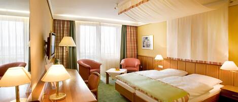 A hotel room with a bed, two chairs, a desk with a lamp, a TV, and a window with curtains.