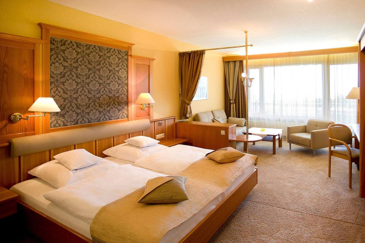 Une chambre d’hôtel avec deux lits, un canapé, un fauteuil, une petite table et une vue sur la ville grâce à de grandes fenêtres.