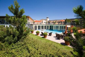 Un área de piscina con mobiliario exterior, rodeada de plantas en macetas y un edificio con balcón.