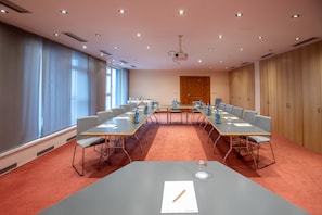 Un salón de conferencias con una larga mesa, sillas, un proyector y un amplio ventanal con cortinas.