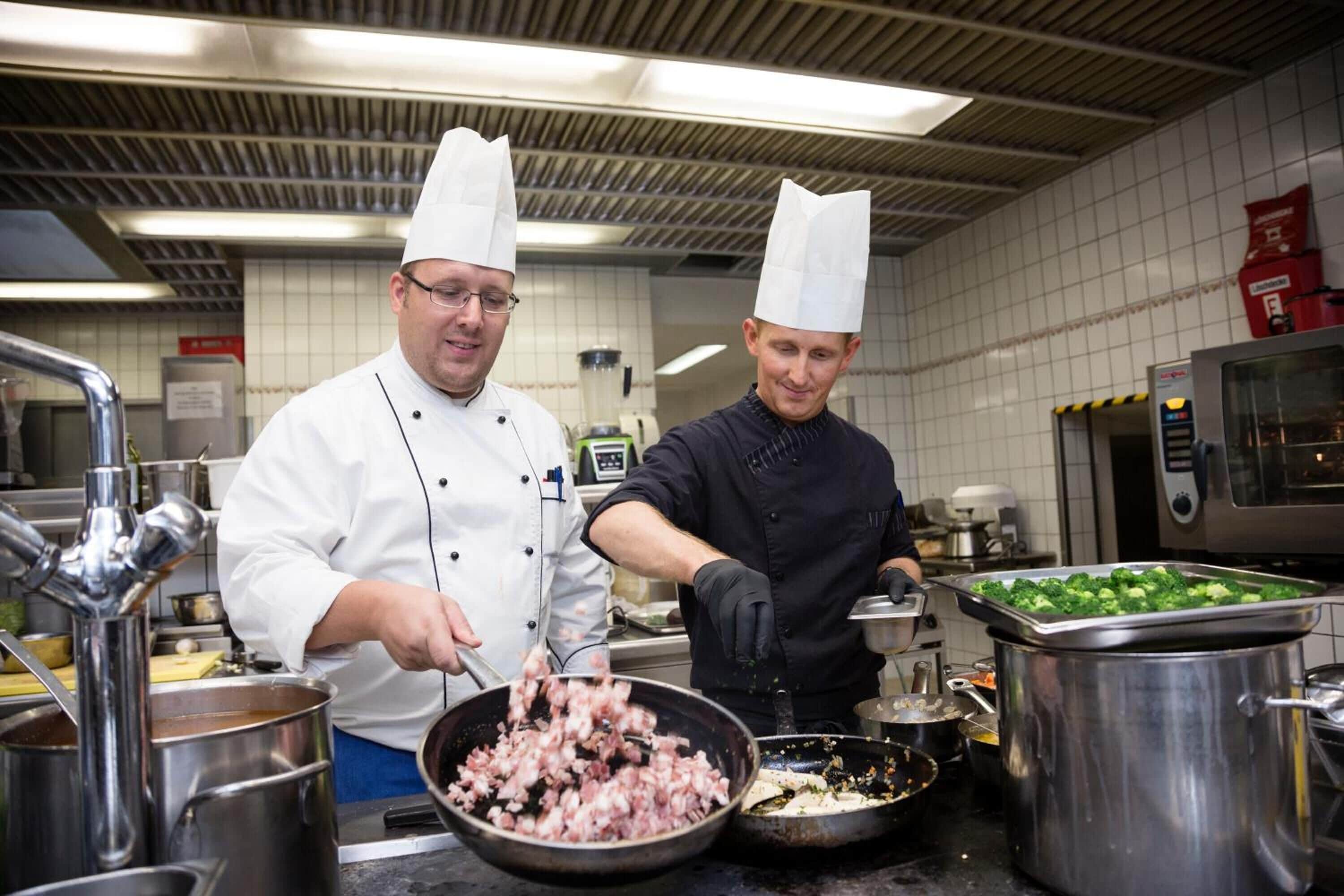 Two chefs in a kitchen with stainless steel equipment and a tiled backsplash.