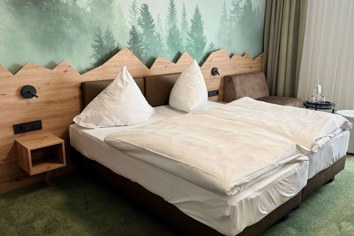 Une chambre d’hôtel avec un grand lit, une tête de lit ornée d’une fresque représentant une forêt, une petite table de chevet et une moquette verte.