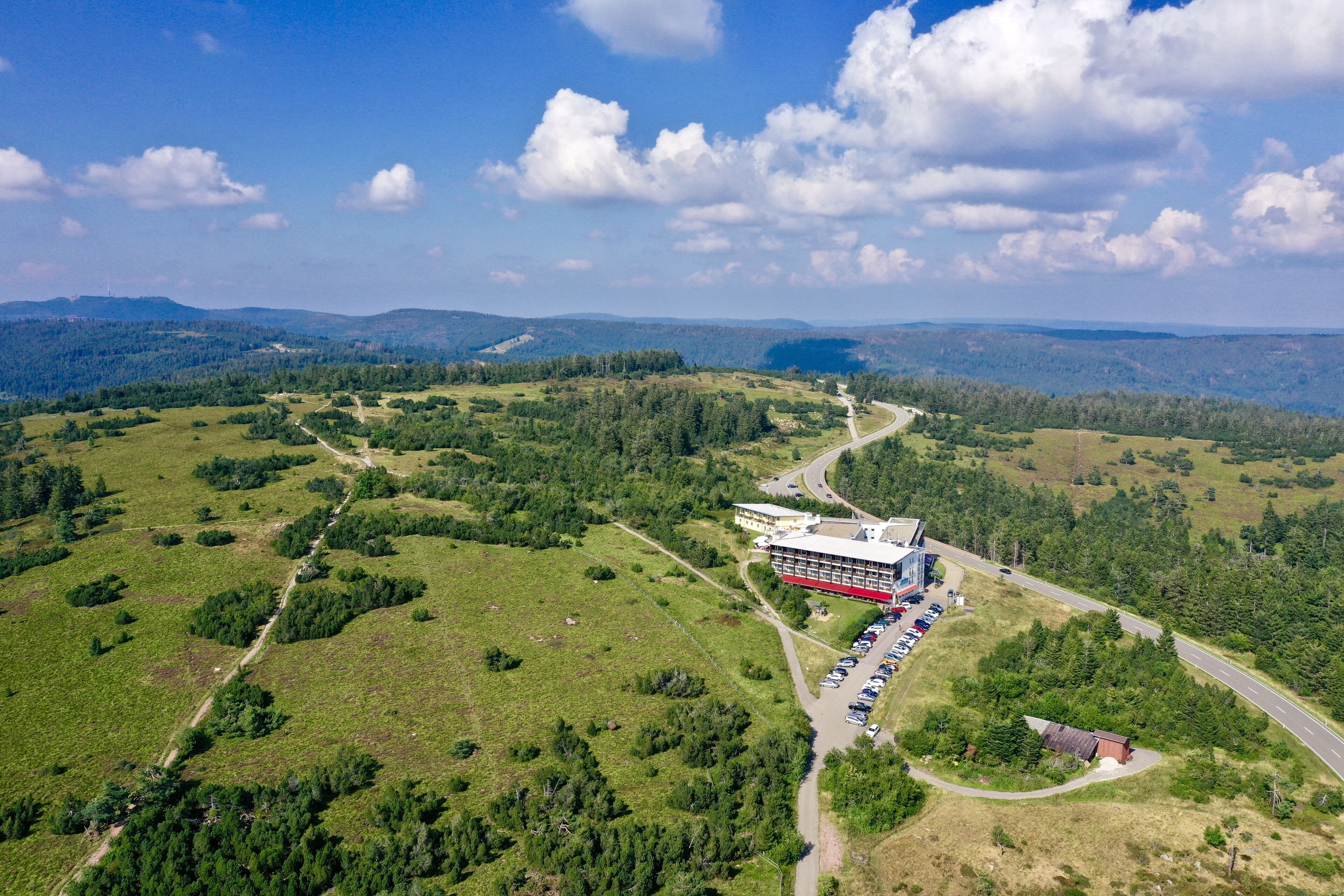 An aerial view of a hotel with a parking lot, surrounded by green fields and trees, with a clear sky.