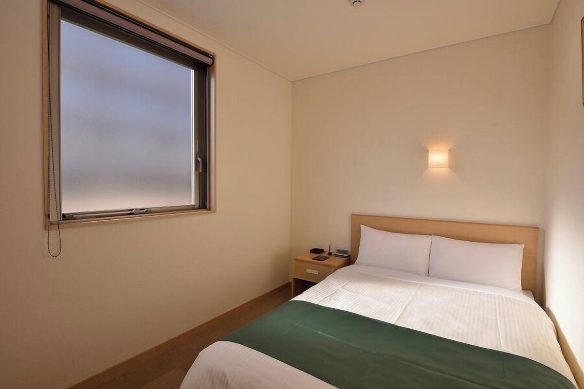 A hotel room with a bed, a bedside table, a nightstand, a window, and a wall-mounted light.