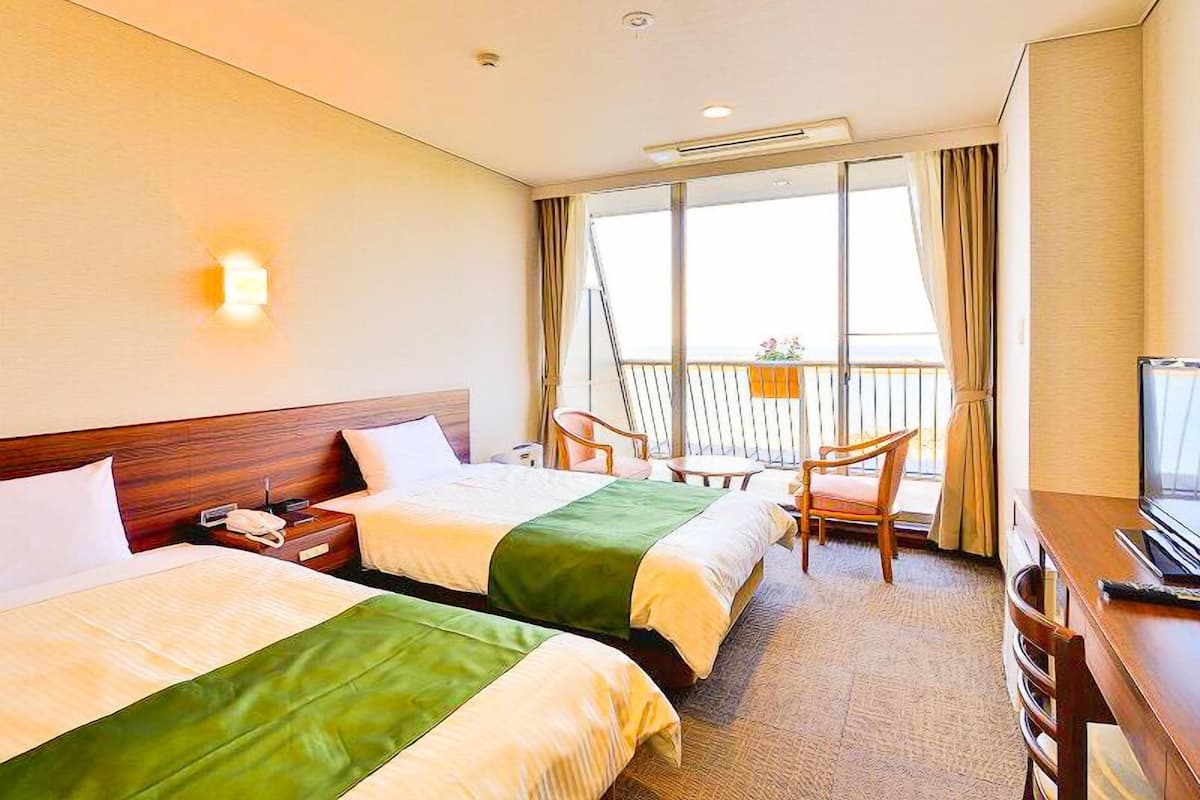 A hotel room with two beds, a desk, a chair, a TV, and a balcony with a view.