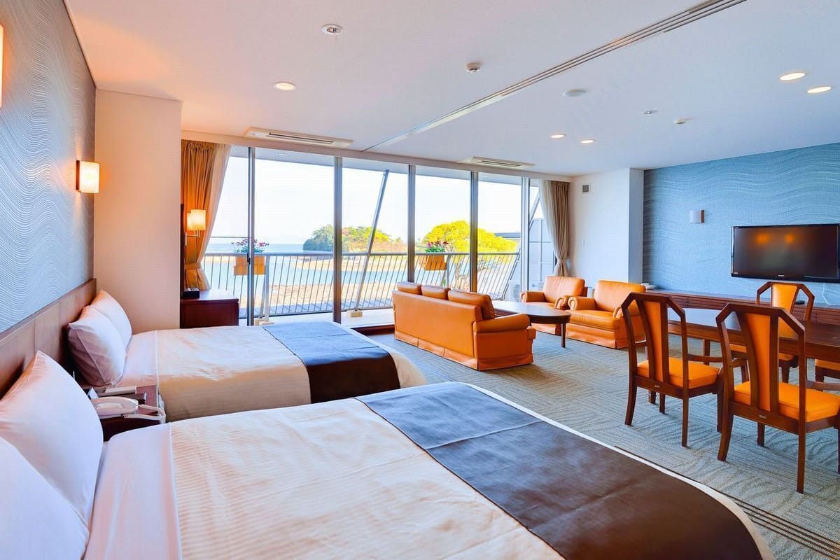 A hotel room with two beds, a sofa, a dining table with chairs, a TV, and a balcony with a view.