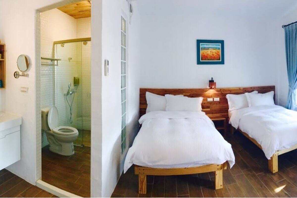 A bedroom with two beds, a wooden headboard, a bedside table, a lamp, and a window with curtains.