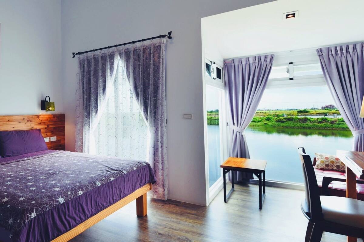 A bedroom with a wooden headboard, a bed with purple bedding, a small table with a chair, and a view of a body of water through the window.