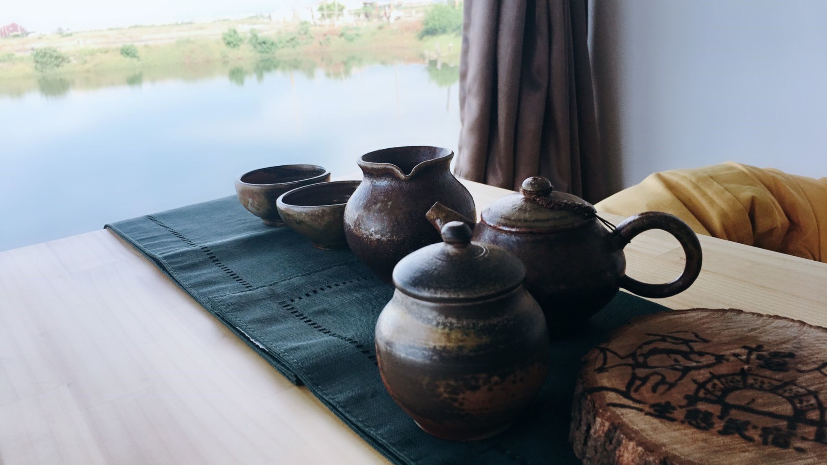Une table ornée de poteries traditionnelles et d’un tapis tissé, un bloc de bois sculpté, et une vue sur un lac paisible, visible à travers une fenêtre.