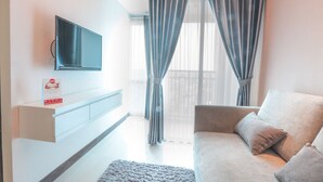 A modern living room with a flat-screen TV mounted on the wall, a sofa with cushions, and a window with curtains. - OYO Flagship 755 Appartel Grand Dhika City (Bekasi)