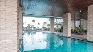 A swimming pool with clear blue water, surrounded by brick walls and columns, with a view of a playground in the background. - OYO Flagship 755 Appartel Grand Dhika City (Bekasi)