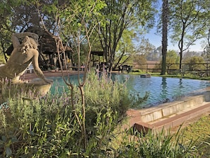 A pool area with a stone sculpture, surrounded by greenery and trees. - Hills and Dales Accommodation (Lanseria)