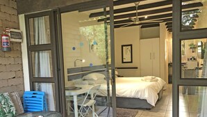 A hotel room with a bed, a small table with chairs, a glass door leading to another room, and a fire extinguisher on the wall. - Hills and Dales Accommodation (Lanseria)